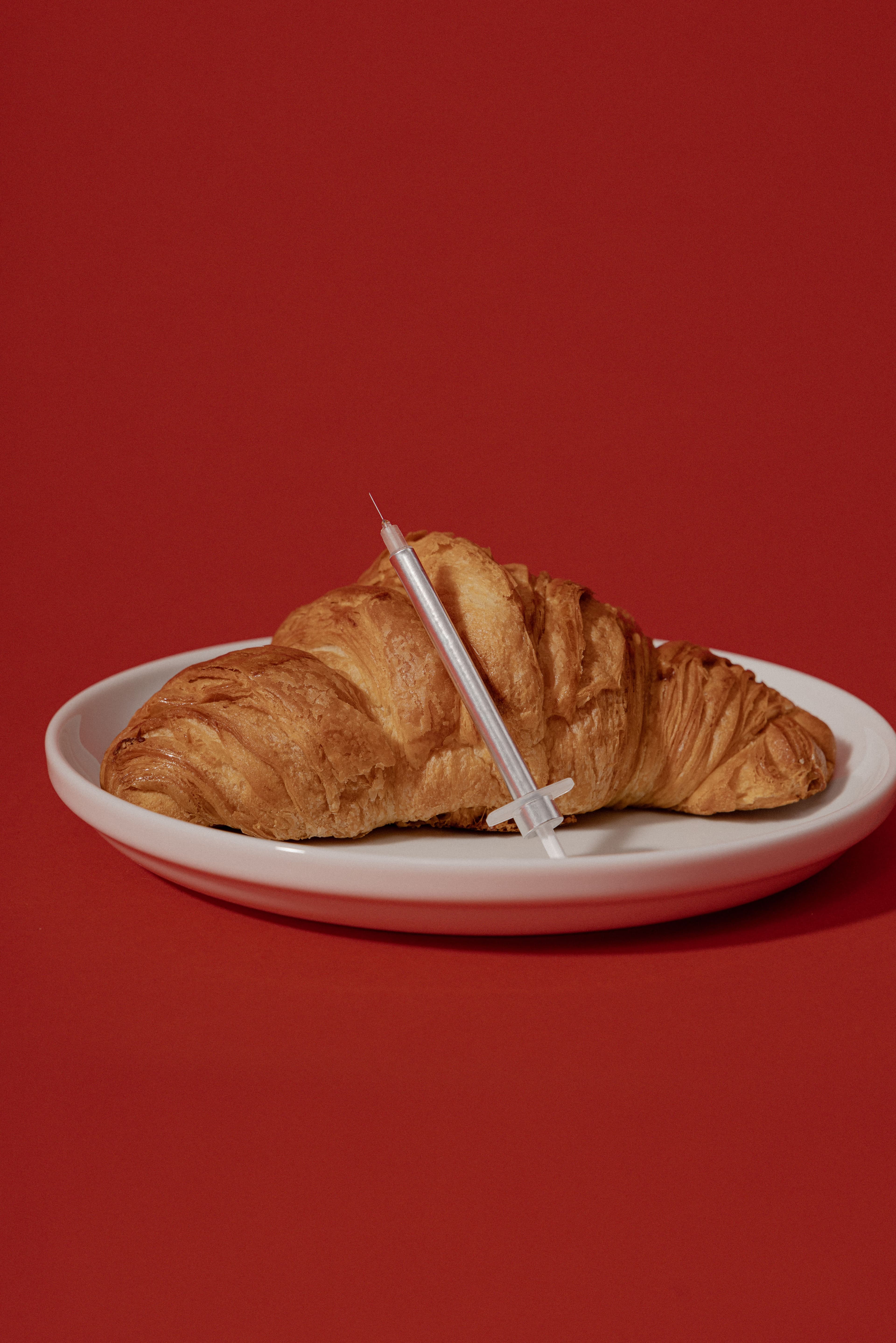 Croissant with silver syringe sitting on a plate in front of a red backdrop.