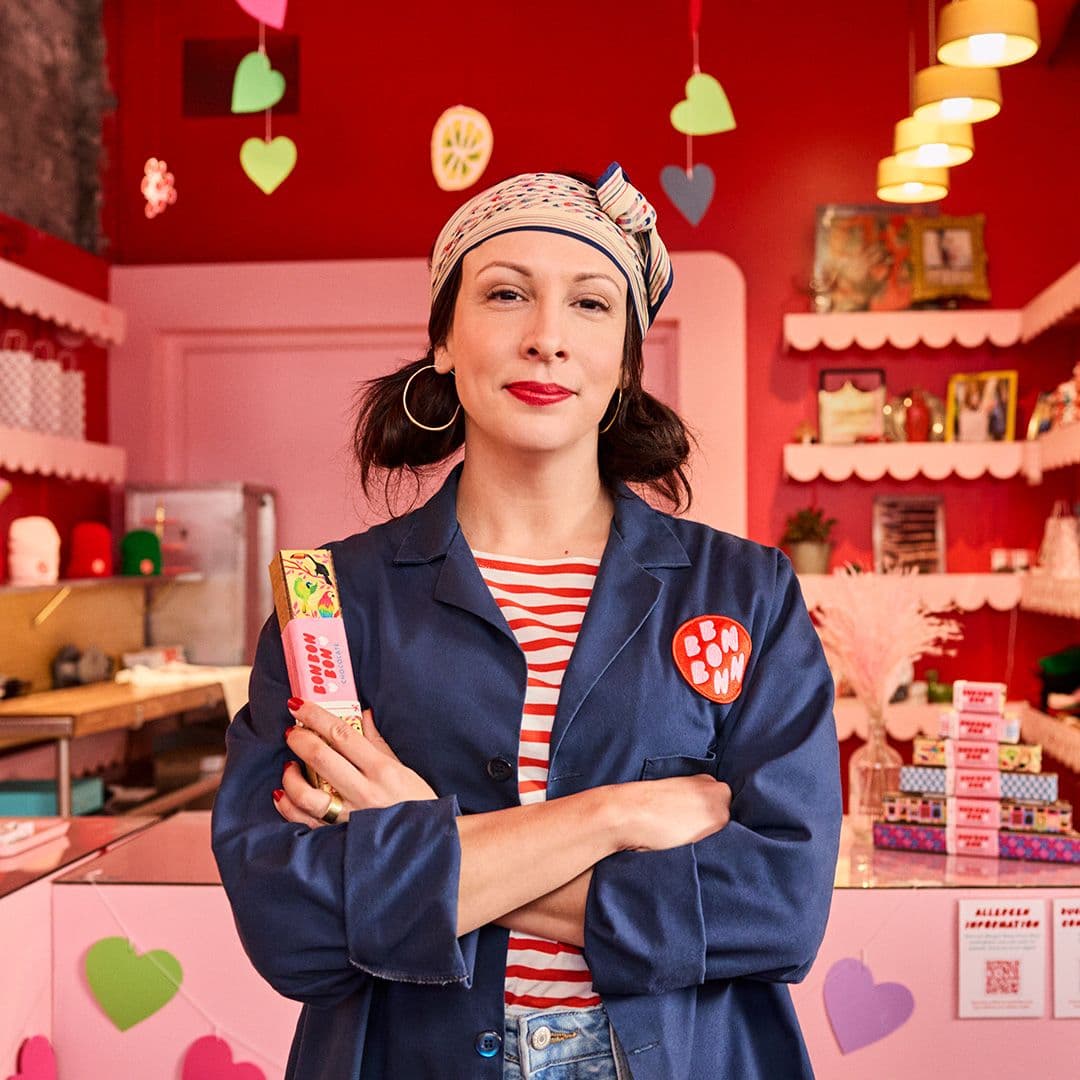 A woman standing in front of a chocolate store with her arms folded across her arms looking proud.